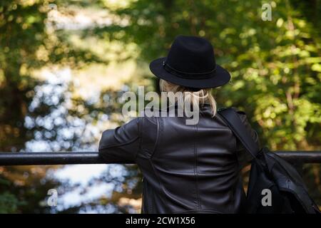 Giovane caucasica in cappello si erge sul ponte e guarda il fiume. Foto Stock