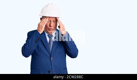 Uomo anziano bello grigio-capelli che indossa vestito e architetto hardhat con mano sulla testa per il dolore in testa perché stress. Che soffre di emicrania. Foto Stock