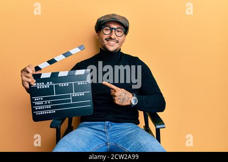 Bell'uomo con tatuaggi che tengono la clapboard del video film seduto sopra la sedia del direttore sorridente punta felice con la mano ed il dito Foto Stock