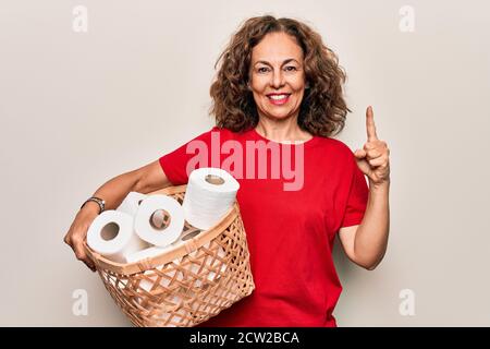 Donna di mezza età che tiene cesto di vimini con un sacco di wc la carta scorre su sfondo bianco sorridendo con un'idea o. domanda che punta il dito con hap Foto Stock