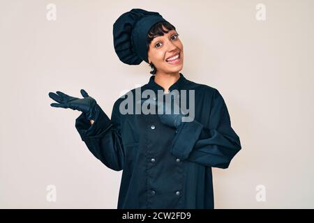 Bella donna brunette con uniforme professionale cuoco stupito e sorridente alla macchina fotografica mentre si presenta con la mano e puntando con il dito. Foto Stock