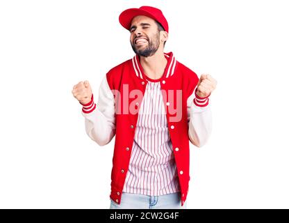 Giovane bell'uomo con barba che indossa giacca da baseball e berretto molto felice ed eccitato facendo gesto vincitore con braccia sollevate, sorridenti e gridanti per Foto Stock