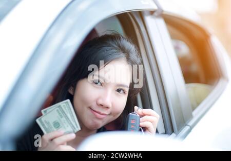 Closeup ritratto felice sorridente attraente donna asiatica seduta nella sua nuova auto bianca mostrando le chiavi, tenendo il dollaro trasporto personale conce acquisto Foto Stock