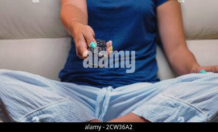 Primo piano del controller nella mano della donna mentre si siede sul divano. Telecomando nelle mani della persona che punta al televisore, premendo il pulsante e cambiando i canali davanti al televisore. Foto Stock