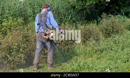 Decespugliatore a benzina. Uomo in tute, occhiali protettivi, cuffie insonorizzate e guanti da lavoro falciano l'erba con taglierina a gas. Vista posteriore completa. Foto Stock