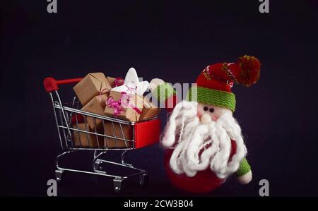 Simpatico giocattolo di Babbo Natale, seduto vicino con carrello rosso. Vendita di Natale o concetto di Capodanno, isolato su sfondo nero Foto Stock