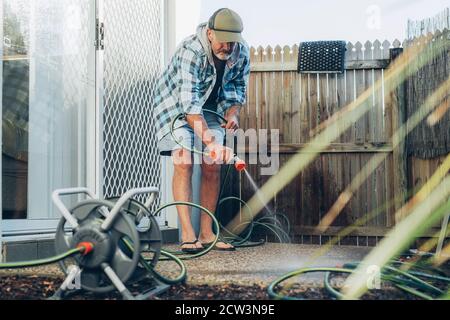 Adulto caucasico medio invecchiato uomo bearded pulisce la terrazza esterna con flusso d'acqua dal tubo flessibile Foto Stock