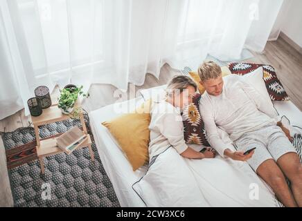 Vista dall'alto della coppia che si trova in un comodo letto in camera da letto e navigare insieme a Internet o controllare le e-mail utilizzando uno smartphone moderno. Tecnologia moderna, e. Foto Stock