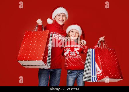 Cute bambini in rosso Santa cappelli, maglioni che tengono borse shopping su sfondo rosso. Foto Stock