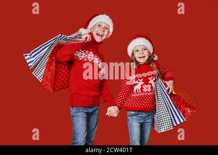 Cute bambini in rosso Santa cappelli, maglioni che tengono borse shopping su sfondo rosso. Foto Stock