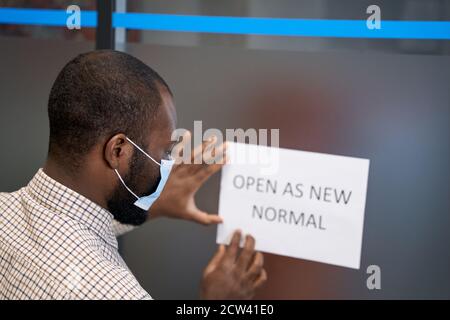 Vista posteriore di un giovane africano che indossa una maschera protettiva Segno aperto bloccato con testo APERTO COME NUOVO NORMALE attivo la porta di vetro in ufficio Foto Stock