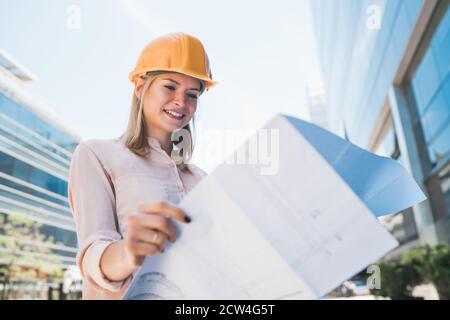 Ritratto di architetto professionista che indossa un casco giallo e guarda le stampe blu all'esterno dell'edificio moderno. Progettazione e architettura. Foto Stock