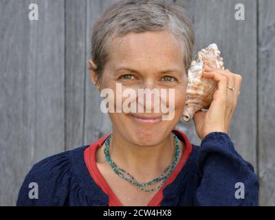 Moderna donna caucasica canadese di mezza età con capelli corti ascolta una conchiglia di mare. Foto Stock