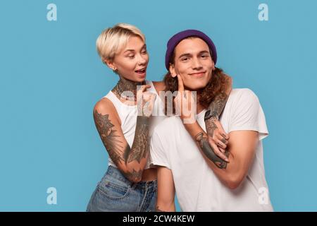 Stile libero. Giovane coppia tatuata in piedi isolata su ragazza blu abbracciando il ragazzo chiedendo di baciarlo gioioso Foto Stock
