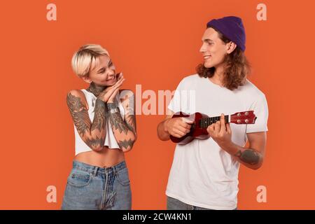 Stile libero. Giovane coppia tatuata in piedi isolato su ragazza arancione guardando ragazzo toccato mentre lui giocare ukulele sorridendo fiducioso Foto Stock