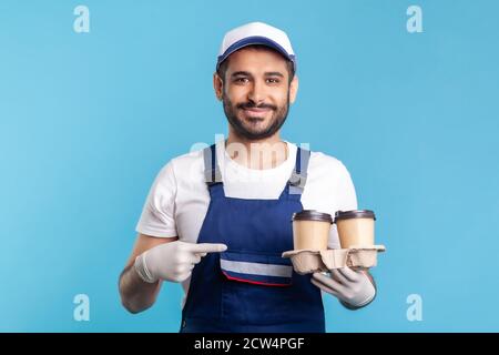 Servizio di consegna. Uomo di corriere ottimista in tute che punta al caffè, indossando guanti di sicurezza che offrono bevande in tazze usa e getta e guardando camer Foto Stock