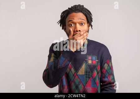 Uomo africano impaurito con la bocca di chiusura temadlock con la mano guardando la macchina fotografica con gli occhi grandi, mantenendo segreto, scioccato si chiedeva con le notizie. Monolocali interni Foto Stock