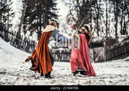 Battaglia di due cavalieri in abiti colorati e cappelli vicino alla foresta e fortezza in legno. I cavalieri combattono in inverno, nella neve. Foto Stock