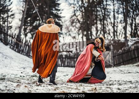 Battaglia di due cavalieri in abiti colorati e cappelli vicino alla foresta e fortezza in legno. I cavalieri combattono in inverno, nella neve. Foto Stock