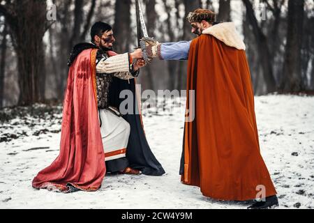 Battaglia di due cavalieri in abiti colorati e cappelli vicino alla foresta e fortezza in legno. I cavalieri combattono in inverno, nella neve. Foto Stock