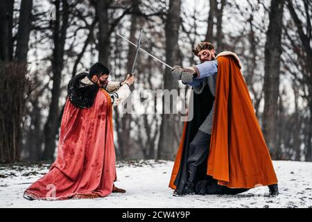 Battaglia di due cavalieri in abiti colorati e cappelli vicino alla foresta e fortezza in legno. I cavalieri combattono in inverno, nella neve. Foto Stock