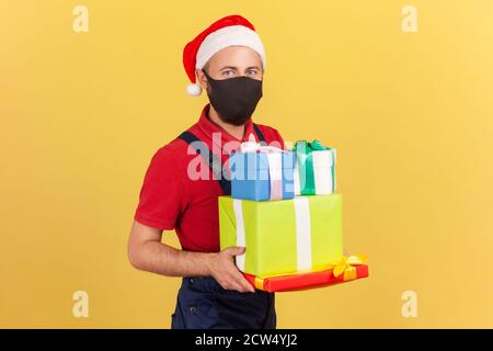 Postino in uniforme maschera protettiva e cappello babbo natale tenendo e mostrando pacchi e regali, consegna di regali vacanze durante la quarantena. Interno Foto Stock