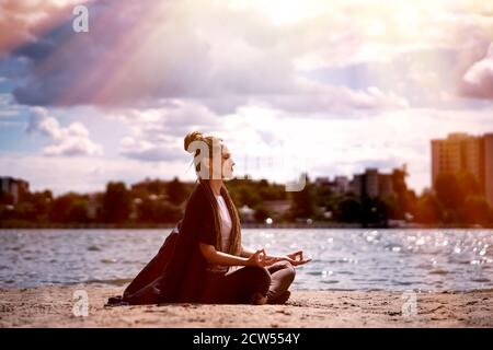 Una giovane donna con una treccia afro in abbigliamento sportivo in una posizione lotus medita sulla spiaggia. Fiume e bellissimo cielo nuvoloso. Concetto di salute e yoga. Foto Stock