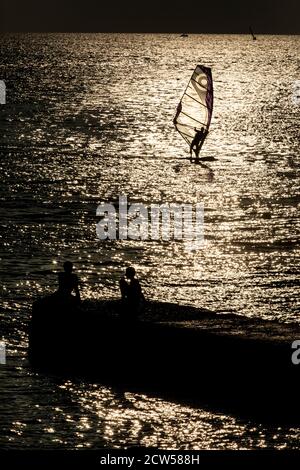 Una silhouette di un windsurfer contro uno sfondo di tramonto sopra una superficie del mare che cattura un vento e un'onda Foto Stock