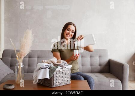 Donna che versa il detergente nel coperchio del flacone. È seduta a tavola con i vestiti raccolti nel cestino Foto Stock