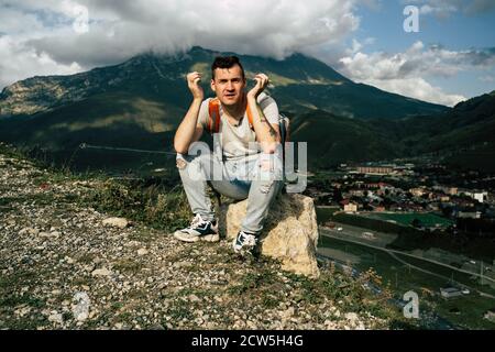 Giovane uomo grimaces, seduto su pietra sullo sfondo del villaggio in montagna. Stanco maschio turista aping, riposante sulla collina dopo trekking attivo in Foto Stock