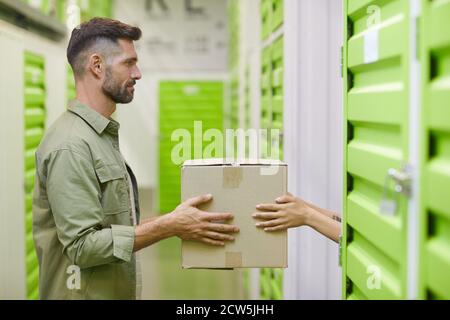 Vista laterale di un bell'uomo portatore che tiene scatola di cartone e che si consegna a donna durante l'imballaggio unità di deposito auto, spazio di copia Foto Stock