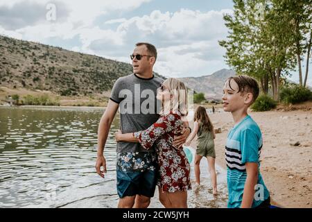 I genitori abbracciano vicino a un lago mentre i loro bambini giocano Foto Stock