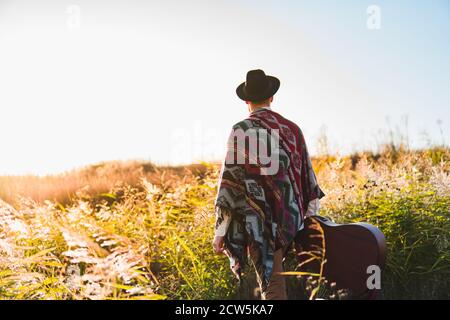 Uomo con chitarra acustica che indossa poncho in una zona rurale al tramonto Foto Stock