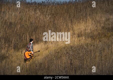 Un musicista di campagna con chitarra acustica cammina nelle erbe a un campo Foto Stock