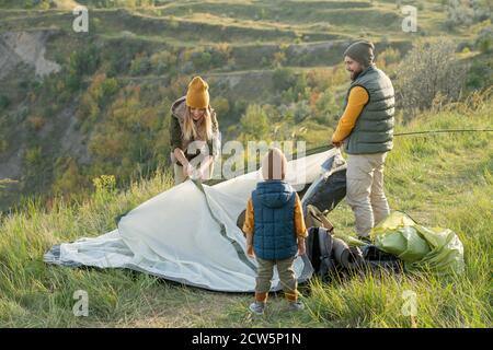 La giovane coppia e il loro figlioletto mettono la tenda sull'erba in cima alla montagna Foto Stock