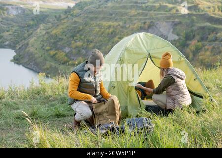 Giovane uomo che guarda in zaino mentre squatting da tenda con sua moglie e suo figlio Foto Stock