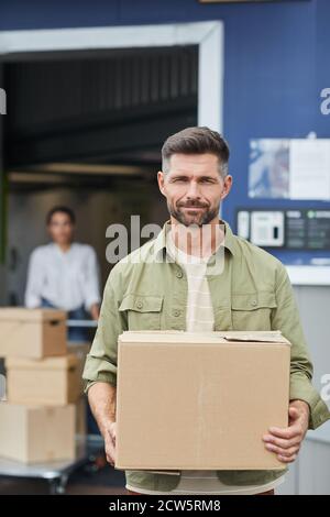 Verticale vita su ritratto del bel uomo che tiene scatola e. guardare la fotocamera mentre si è in piedi da un sistema di deposito autonomo Foto Stock