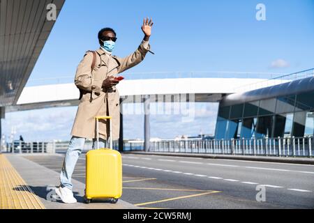 Uomo millenario afro-americano con valigia gialla si trova nel terminal dell'aeroporto, utilizzando il telefono, chiamando il taxi, alzando la mano, indossare maschera medica viso a prote Foto Stock