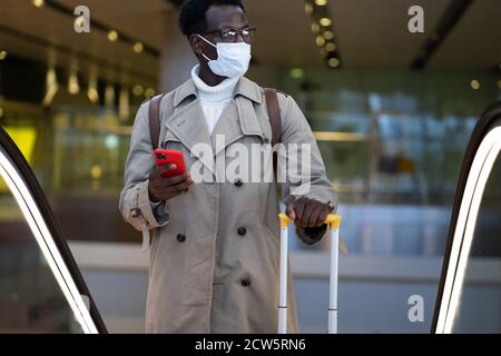 Uomo viaggiatore afro-americano con valigia gialla si trova sulla scala mobile nel terminal dell'aeroporto, indossare maschera medica viso per proteggersi dal contatto con Foto Stock