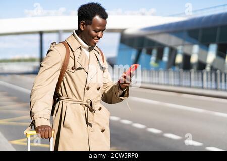 Sorridente uomo afro-americano viaggiatore con valigia si trova nel terminal dell'aeroporto, utilizzando l'applicazione mobile in smartphone per noleggiare un veicolo online in auto sha Foto Stock