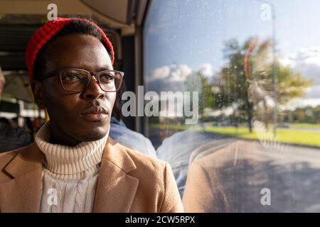 Ritratto dell'uomo viaggiatore afro-americano messo a fuoco in camice beige, maglione bianco e cappello rosso, indossando occhiali, guardando la finestra, viaggiando dal pubblico Foto Stock