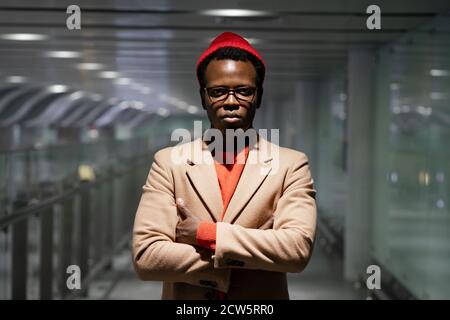 Elegante uomo millenario afro-americano in occhiali indossare cappotto beige, maglione rosso e cappello, in piedi nella sala dell'aeroporto, guardando la fotocamera, le braccia incrociate Foto Stock