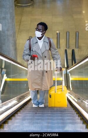 Uomo viaggiatore afro-americano con valigia gialla si trova sulla scala mobile nel terminal dell'aeroporto, indossare maschera medica viso per proteggersi dal contatto con Foto Stock