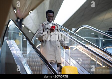 Sorridente viaggiatore afro-americano con valigia gialla si trova sulla scala mobile del terminal dell'aeroporto, utilizzando il telefono cellulare, arriva dall'estero. Foto Stock