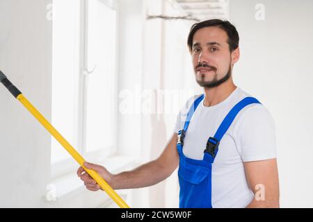 Colpo medio, operaio di costruzione in abiti da lavoro blu dipinge muro in appartamento Foto Stock