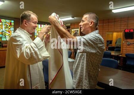 Il diacono di una chiesa cattolica della California meridionale aiuta a vestirsi il suo sacerdote in vestimenti cerimoniali prima della messa. Foto Stock