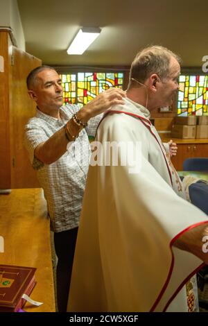 Il diacono di una chiesa cattolica della California meridionale aiuta a vestirsi il suo sacerdote in vestimenti cerimoniali prima della messa. Foto Stock