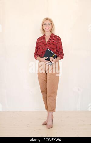 Donna d'affari allegra matura in blusa rossa e pantaloni beige che tengono notebook Foto Stock