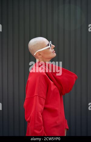 Donna calva cool che indossa felpa con cappuccio rossa e occhiali da sole in piedi su sfondo. Foto Stock