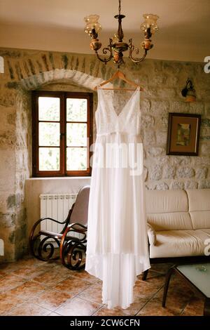 Abito da sposa della sposa su un appendiabiti in legno su un lampadario in una camera con un divano e una sedia a dondolo. Foto Stock